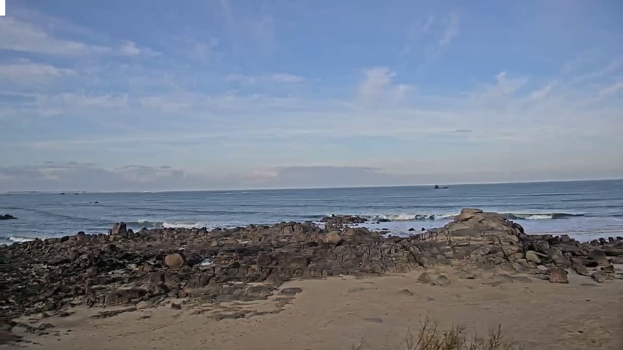 Large waves crash onto a sandy beach and rocky shore under a dark, heavily overcast sky with visible camera lens droplets, suggesting stormy and wet weather along a coastline featuring distant houses and a grassy dune.