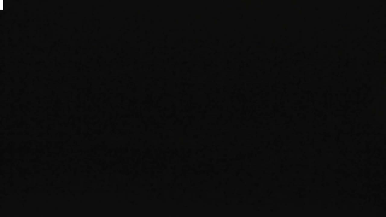 Large waves crash onto a sandy beach and rocky shore under a dark, heavily overcast sky with visible camera lens droplets, suggesting stormy and wet weather along a coastline featuring distant houses and a grassy dune.