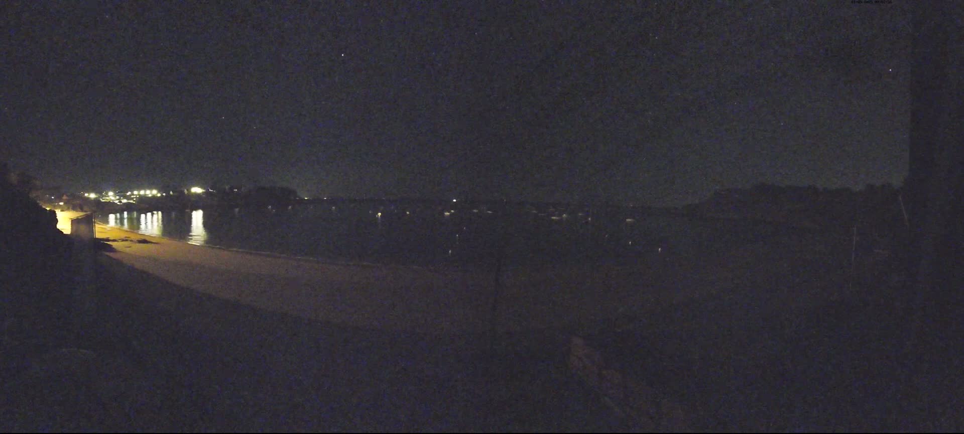 A dark, nighttime view shows a calm body of water reflecting distant lights, a sandy beach, and a shadowed shoreline under a clear, starlit sky.