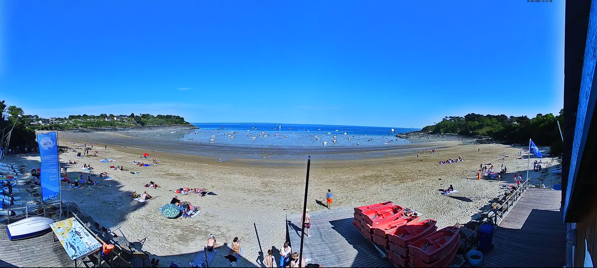 A sandy beach with many people sunbathing and swimming, numerous small boats in shallow water, and a clear blue sky.