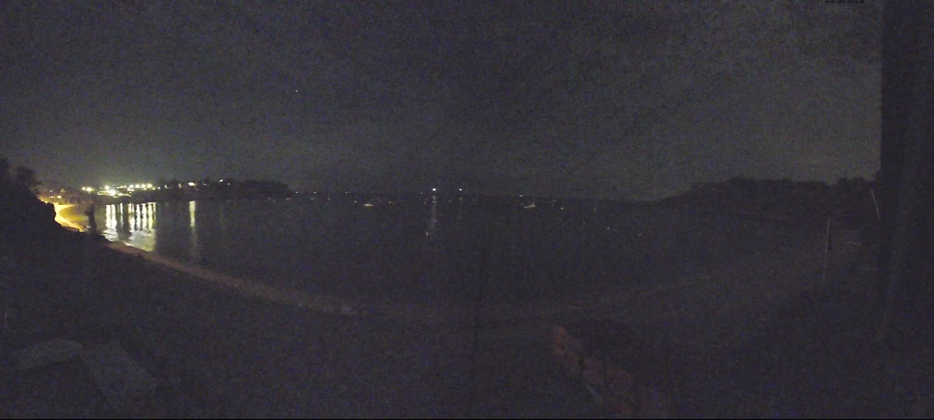 A nighttime view shows a dark body of water reflecting the lights of a distant shoreline under a clear, dark sky.