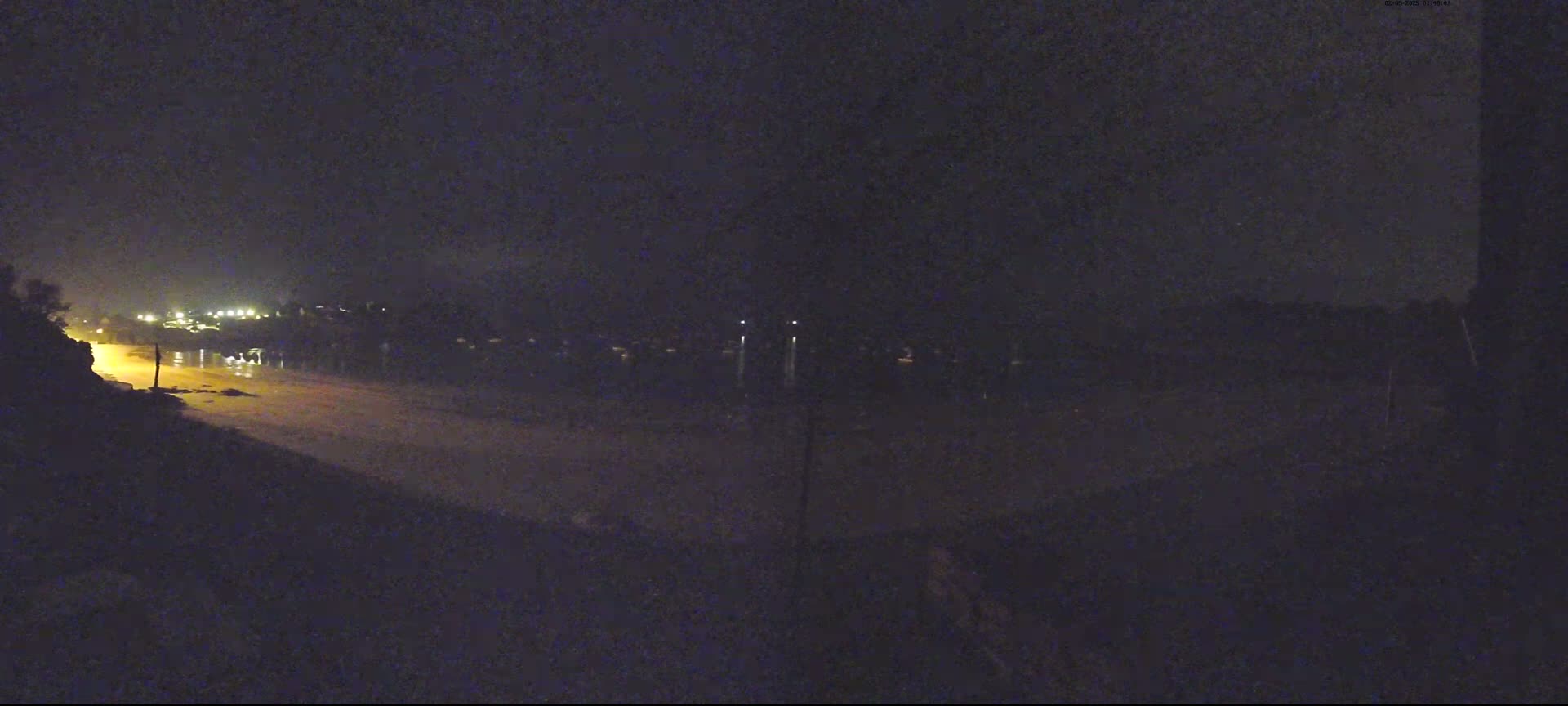A nighttime view of a dark, sandy shoreline with dimly lit buildings and lights across the water under a dark sky.