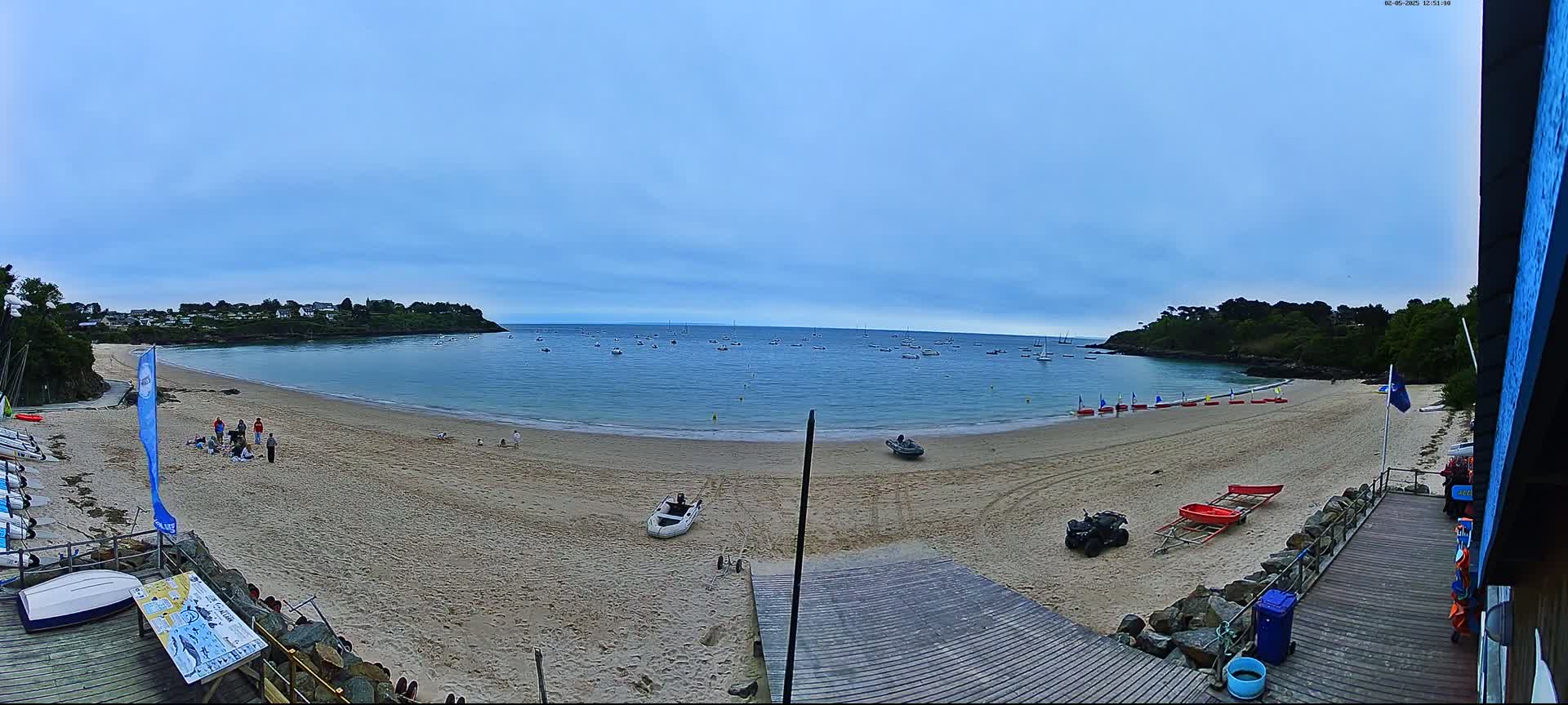 A sandy beach with calm water and numerous boats in the distance under a mostly clear, light-blue sky.