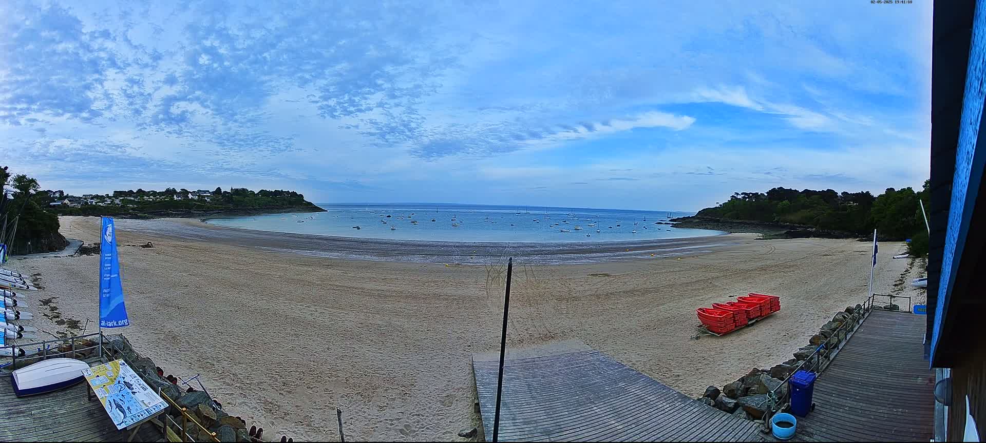 A sandy beach curves around a calm bay dotted with small boats under a partly cloudy sky.