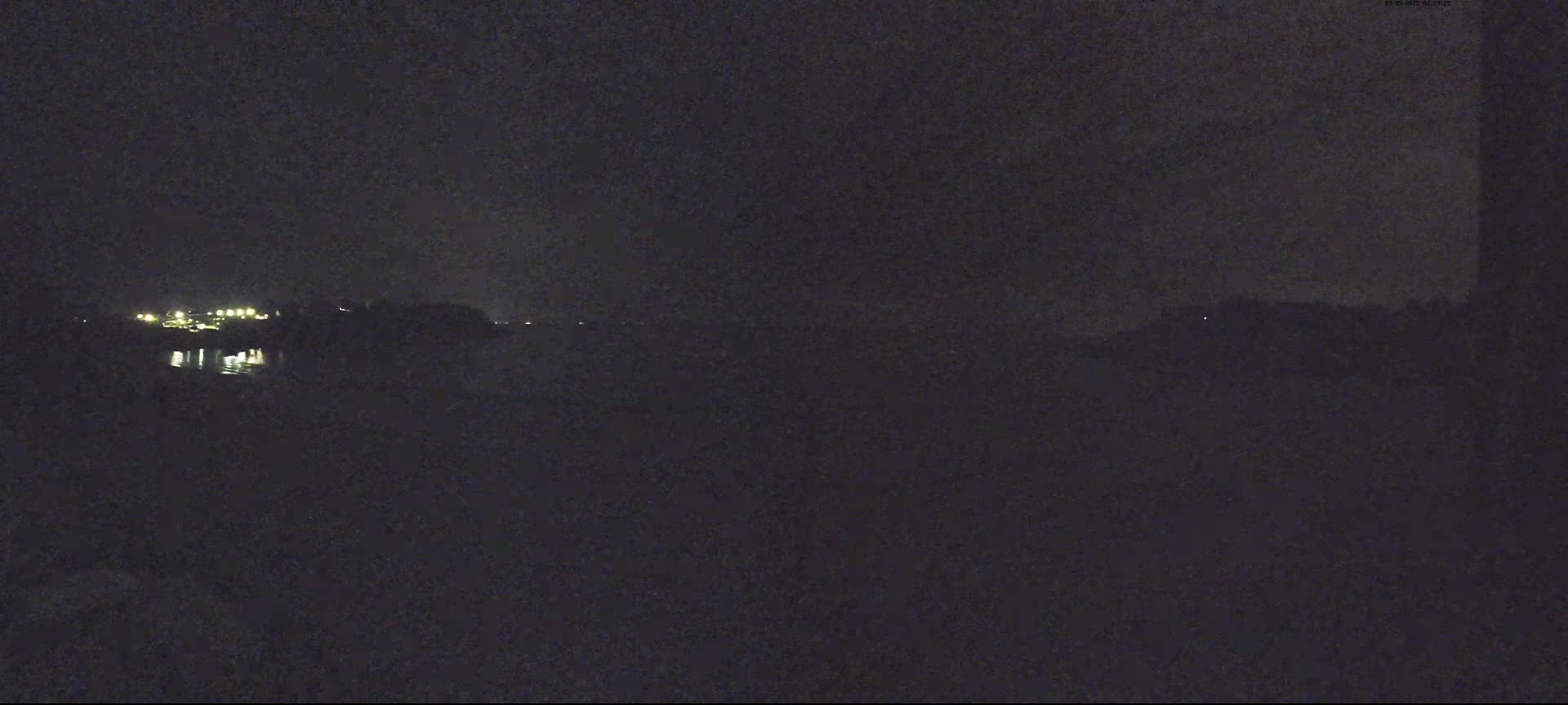 A dark, nighttime scene shows a body of water reflecting the lights of a distant structure across its banks.