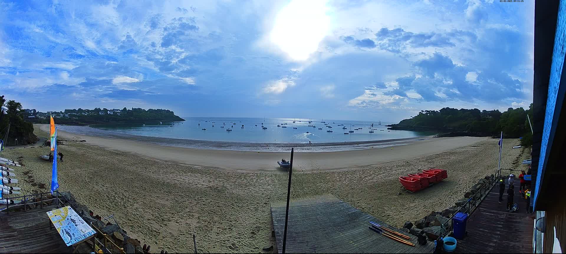 A sandy beach with calm water and numerous boats in the bay under a partly cloudy sky.