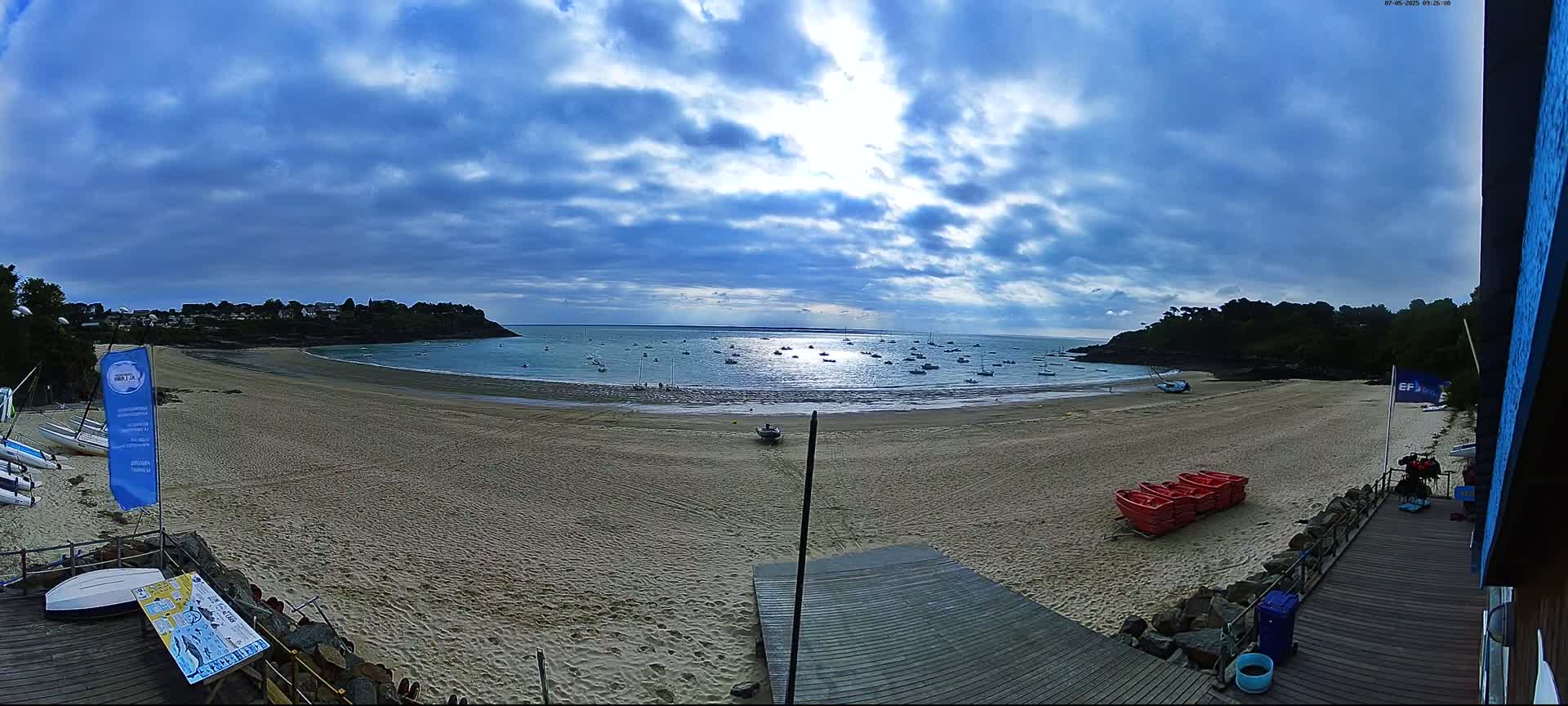A sandy beach with several small boats in the calm water, under a partly cloudy sky, is viewed from a wooden deck near a building.