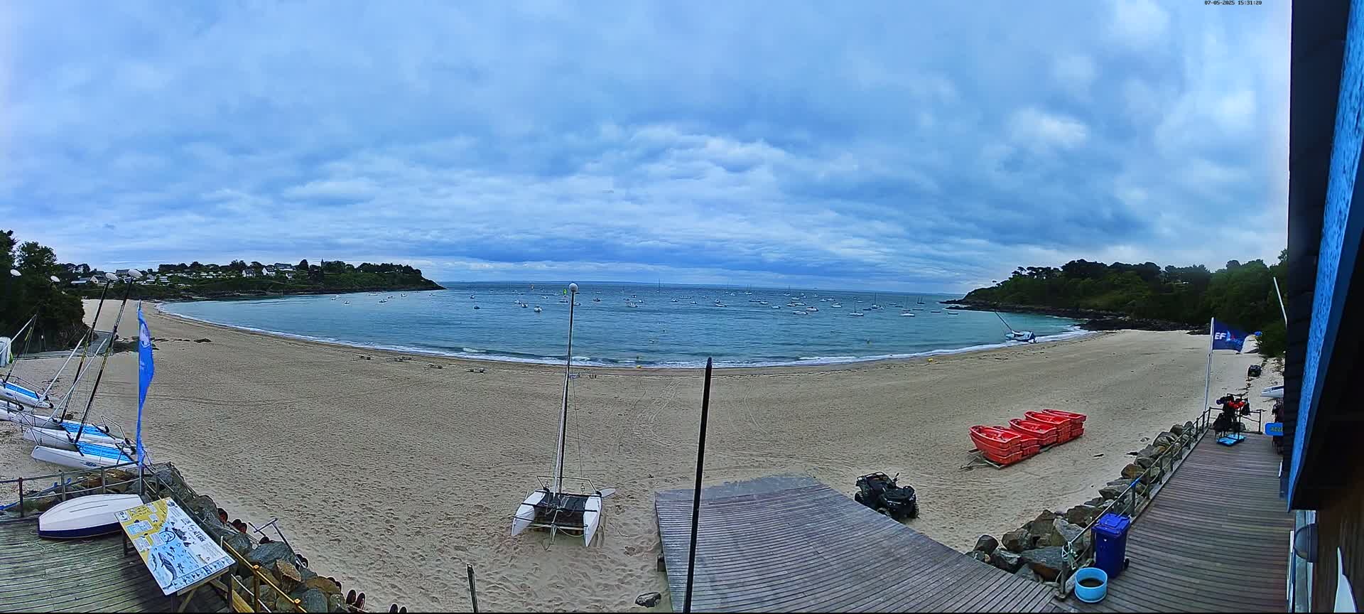 A sandy beach with calm water, numerous small boats in the bay, and several sailboats on the shore under a partly cloudy sky.