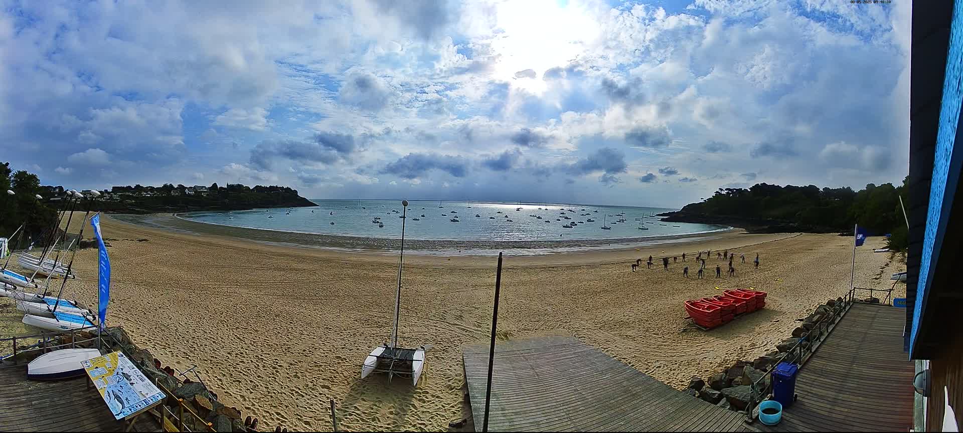 A sandy beach with numerous sailboats in the calm water,  people exercising on the sand, and several small boats are pulled up on the shore under a partly cloudy sky.