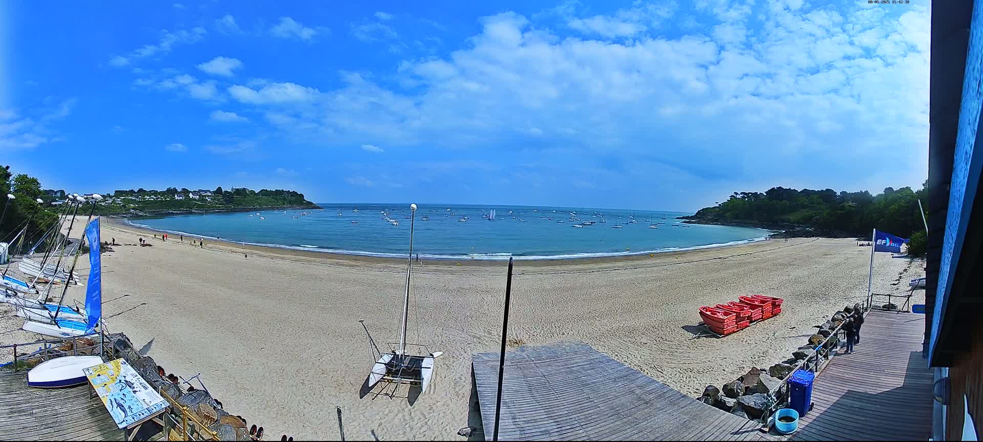 A sandy beach curves around a calm bay dotted with sailboats under a mostly sunny sky.