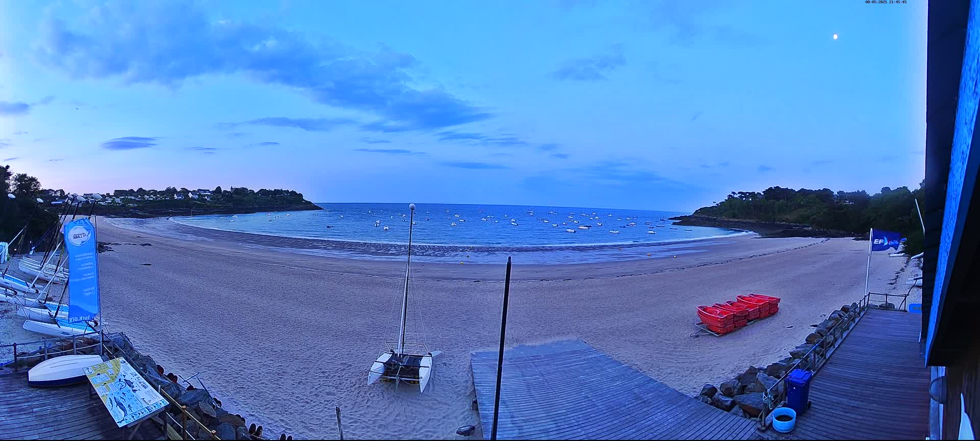 A sandy beach with calm water, numerous small boats in the bay, and several sailboats on shore under a twilight sky.