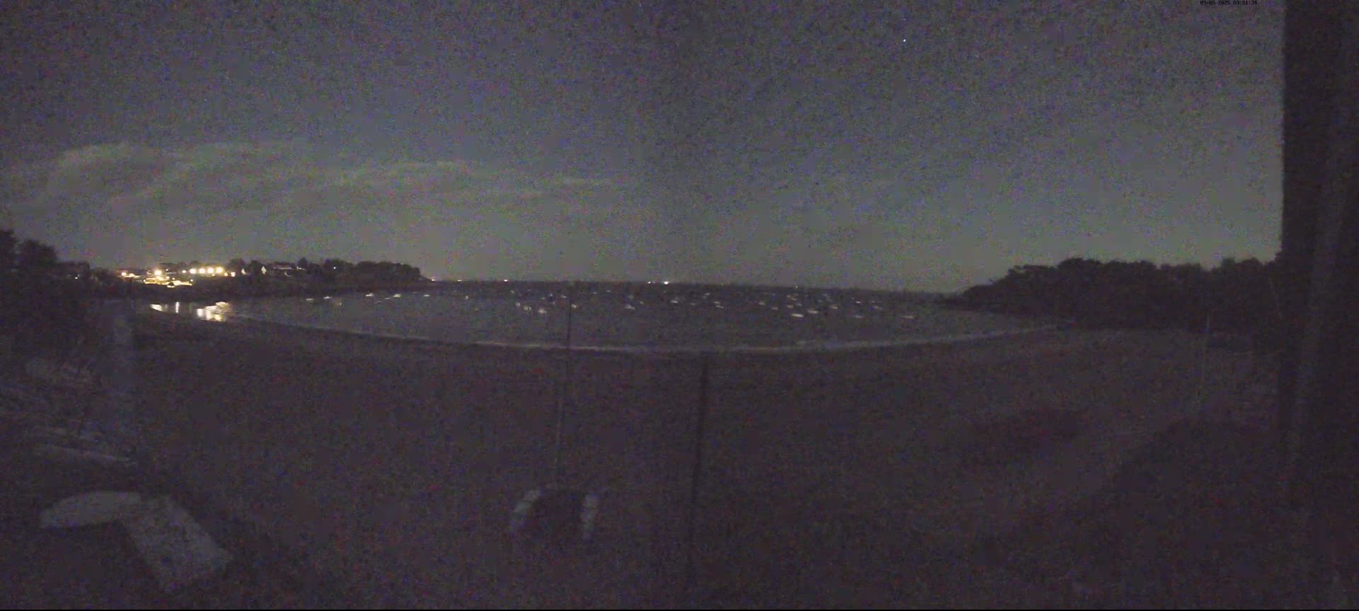 A nighttime view of a dark body of water with a few lights visible on the opposite shore under a mostly clear, dark sky.