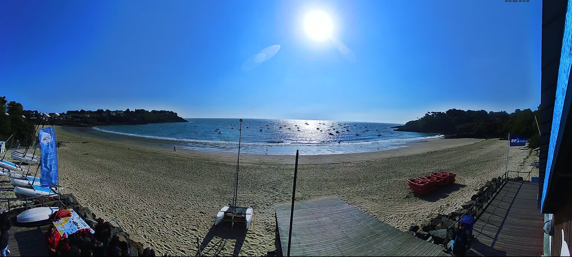 A sandy beach with several sailboats in the water and a bright sun in a clear blue sky.