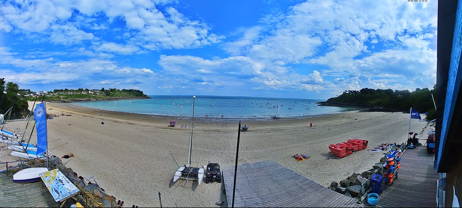 A sandy beach with sailboats in the water under a partly cloudy blue sky.