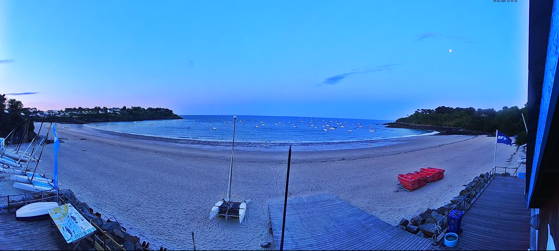A sandy beach at dusk, with calm water and numerous boats at anchor, is viewed from a wooden deck, with several sailboats moored nearby.