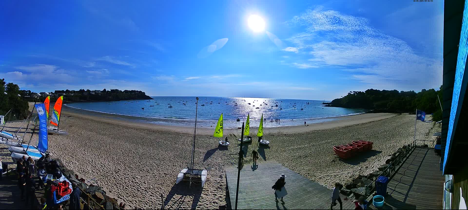 A sandy beach with sailboats in the water under a bright sunny sky, with several people walking on a wooden boardwalk leading to the beach.
