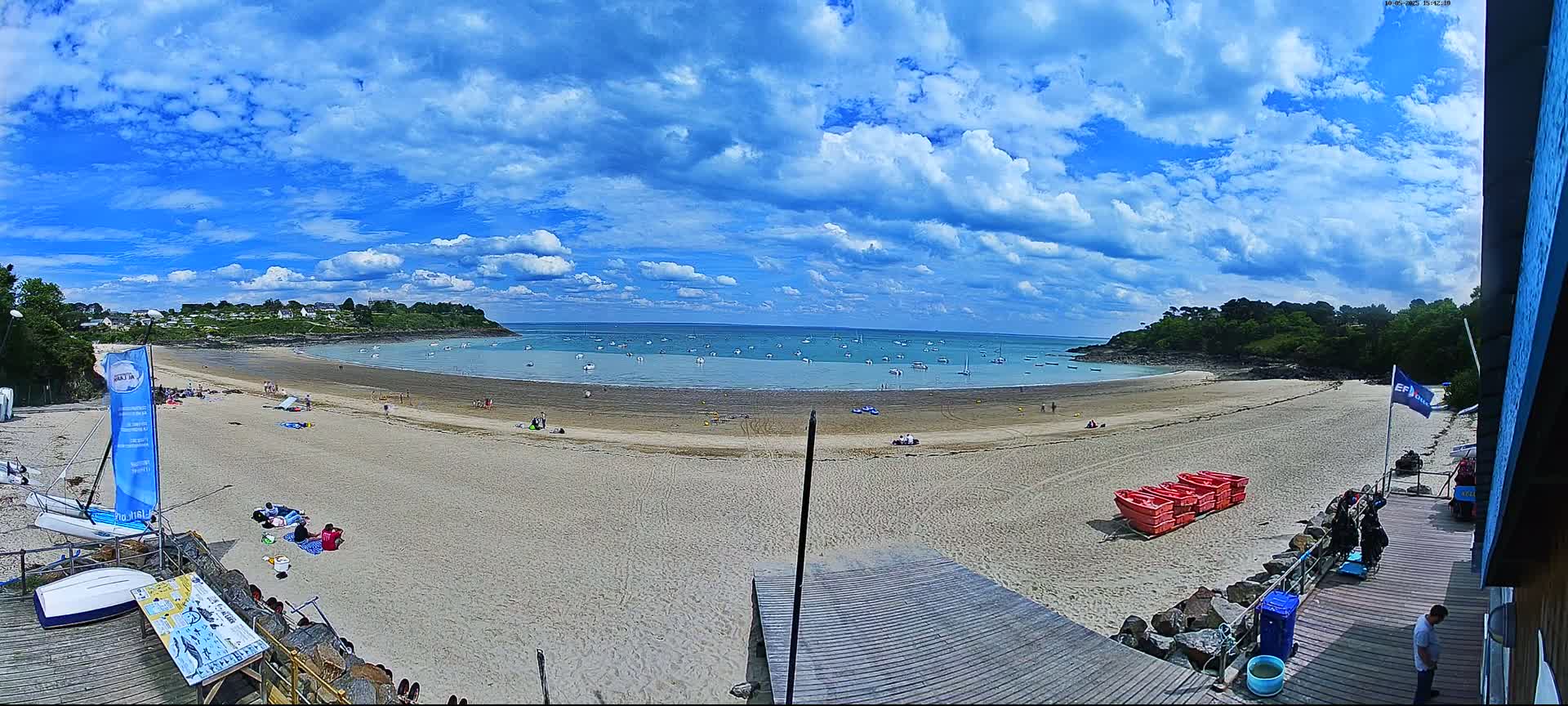A sandy beach curves around a calm bay dotted with sailboats under a partly sunny sky.