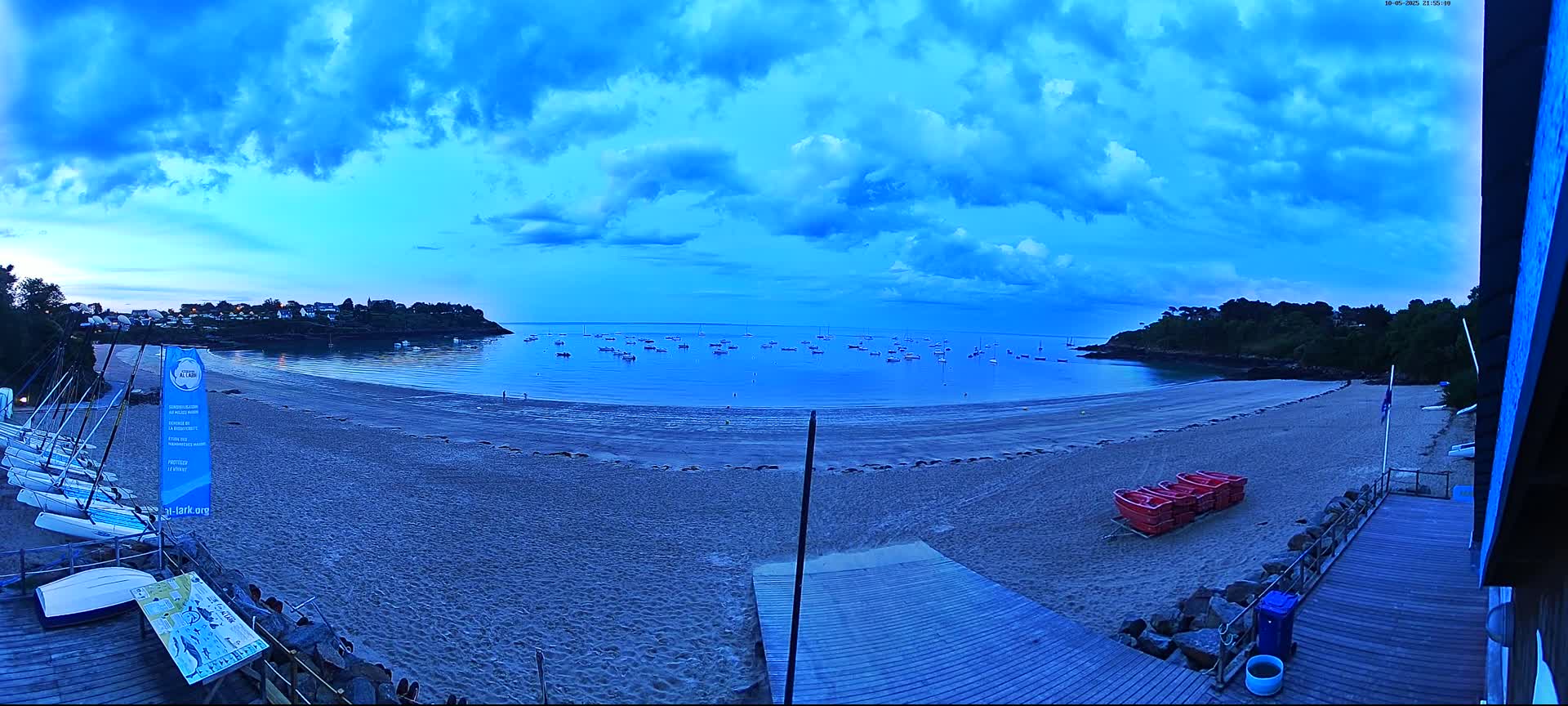 A sandy beach at twilight, under a mostly cloudy blue sky, features numerous small boats in the calm water, several sailboats on the shore, and a wooden boardwalk leading to the beach.