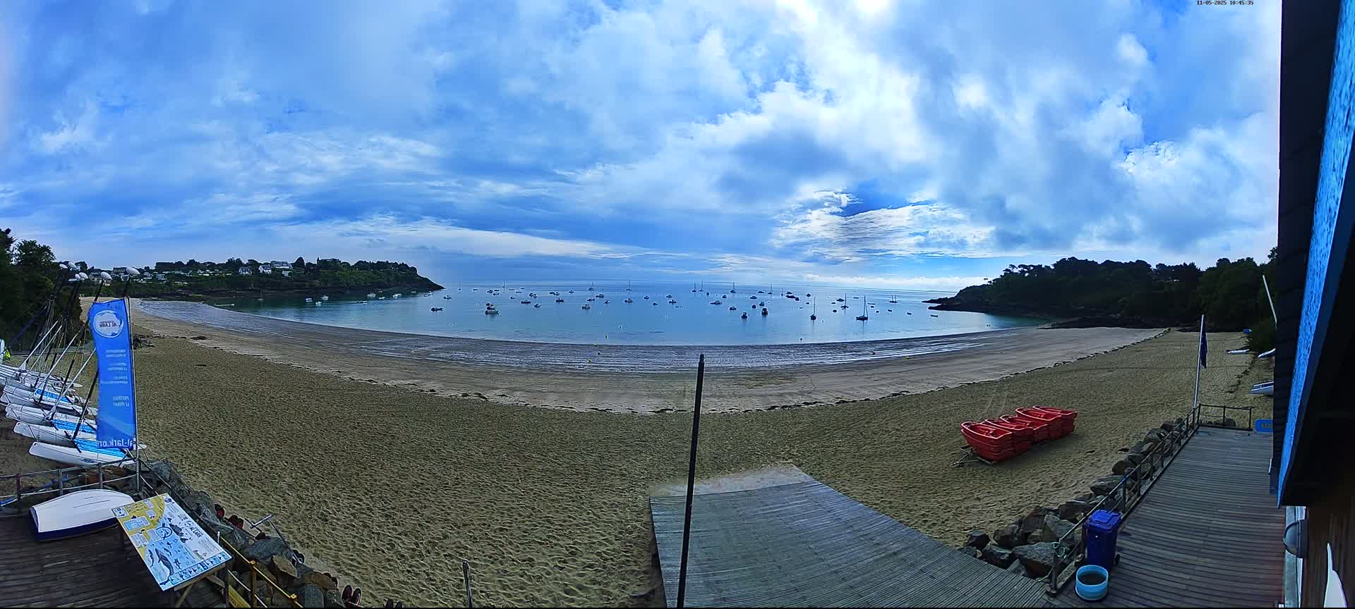 A sandy beach curves around a calm bay dotted with boats under a partly cloudy sky.