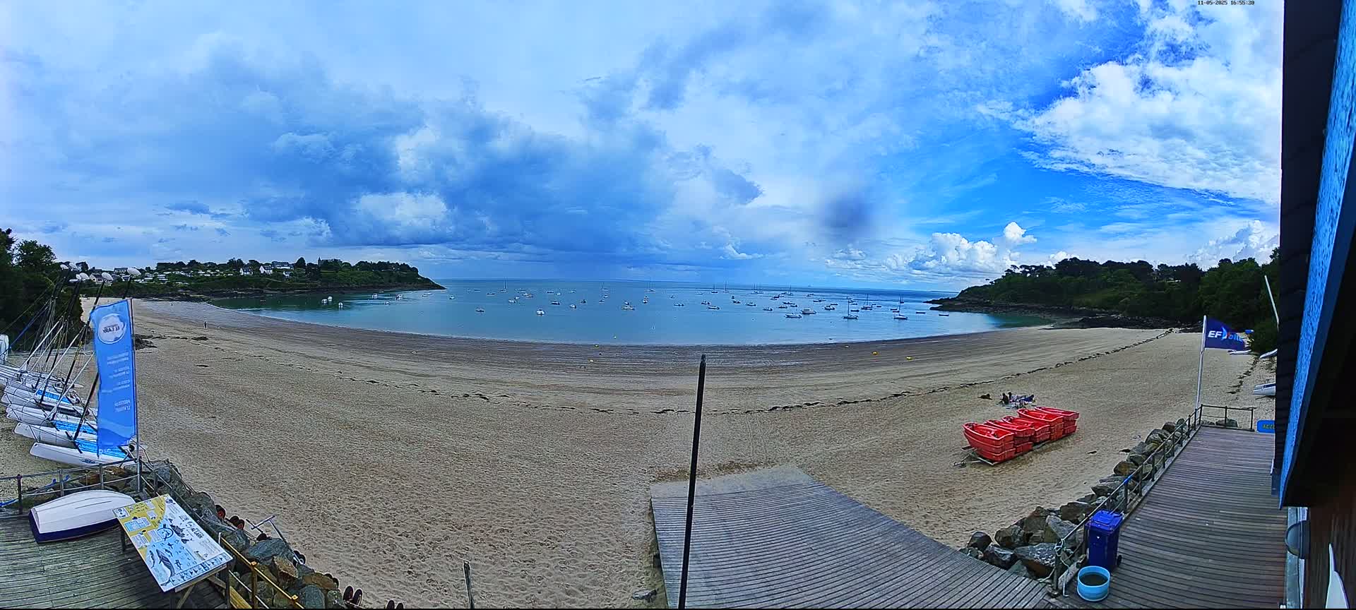 A sandy beach curves around a calm bay dotted with sailboats under a partly cloudy sky.