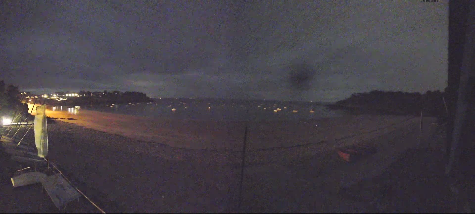 A nighttime view of a calm body of water with several boats moored, a sandy beach, and a dark shoreline under a cloudy sky.