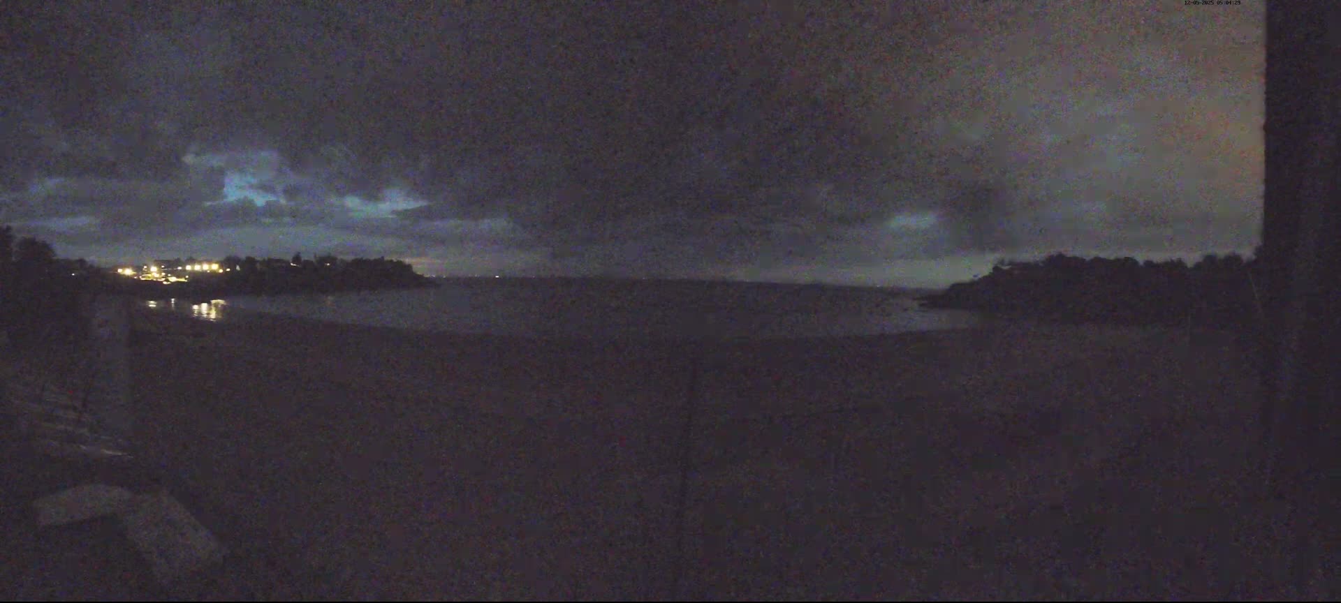 A dark, nighttime view of a body of water with a distant shoreline and faint lights, under a cloudy sky.