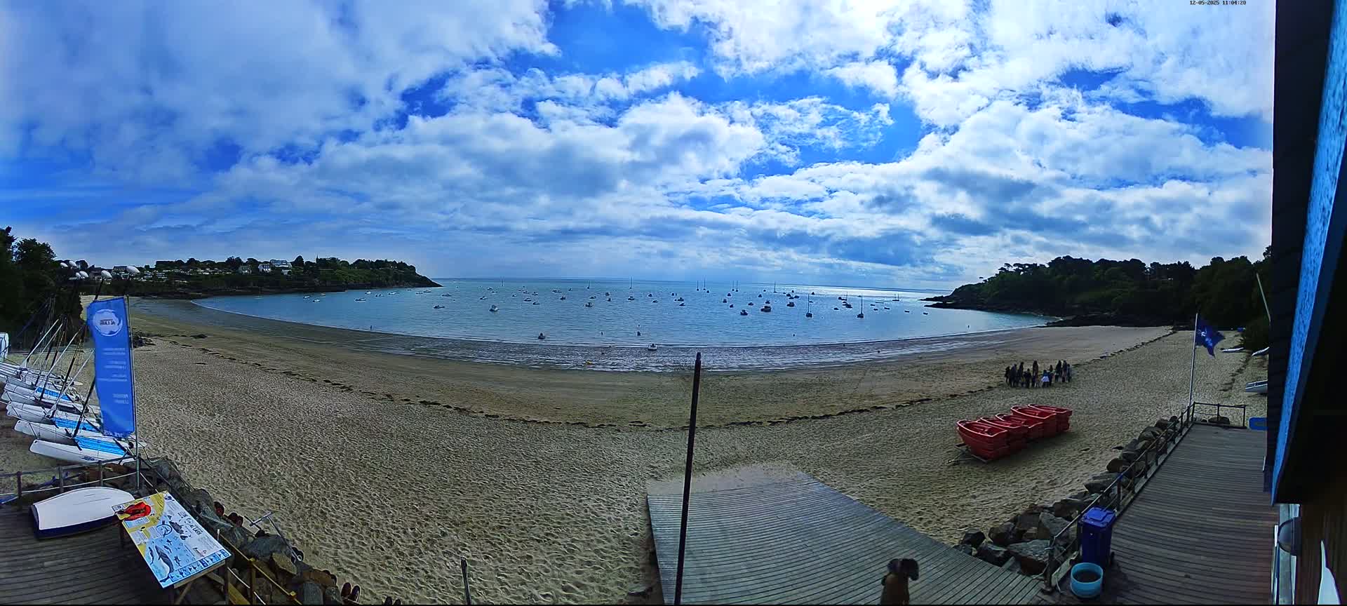 A sandy beach with sailboats in the calm water under a partly cloudy blue sky, with several small red boats and people on the sand.