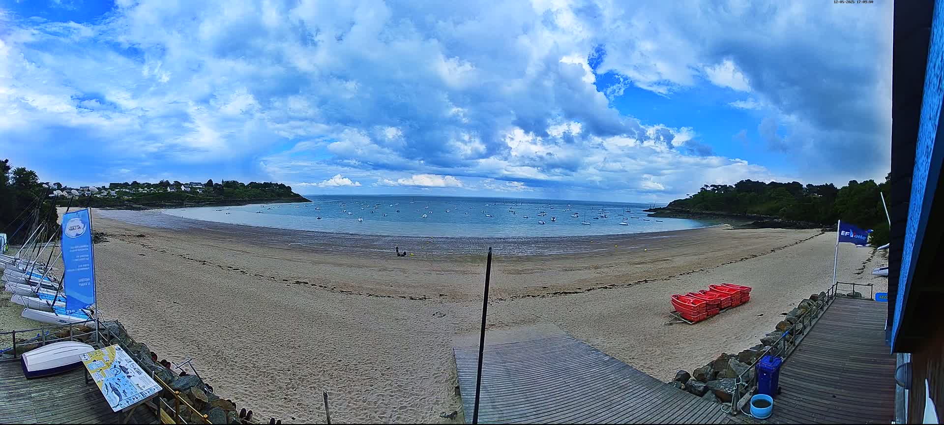 A sandy beach with a calm bay containing numerous small boats is viewed from a wooden boardwalk under a partly cloudy sky.
