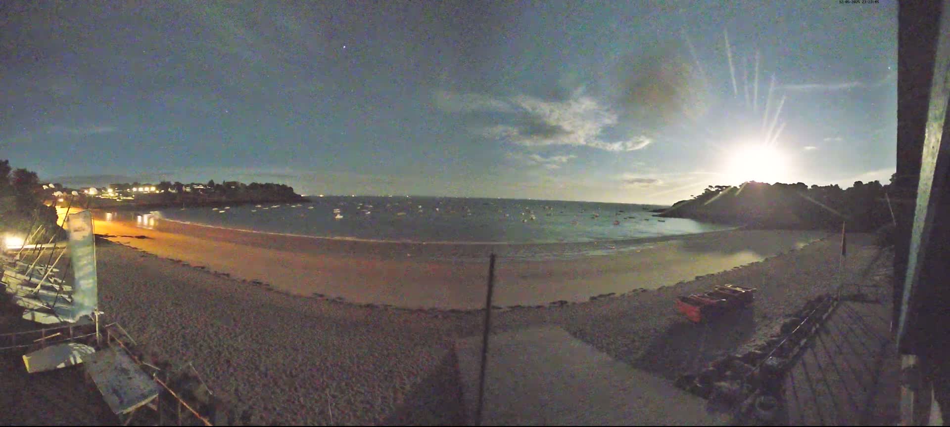 A crescent moon shines over a sandy beach at night, where several small boats are visible on the calm water.