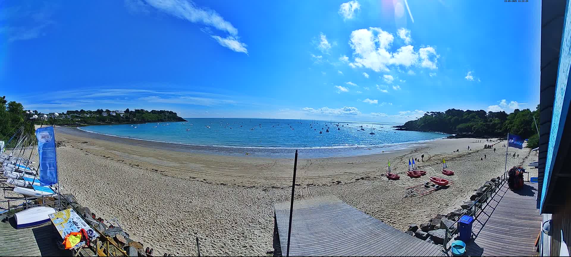 A sandy beach curves around a calm bay under a mostly sunny sky with scattered clouds, boats are visible on the water, and several small sailboats are lined up on the sand near a wooden boardwalk.