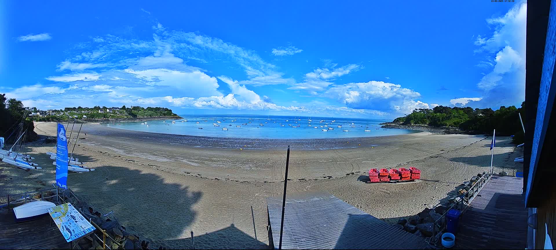 A sandy beach with several sailboats in the calm water, under a mostly sunny sky with puffy white clouds.