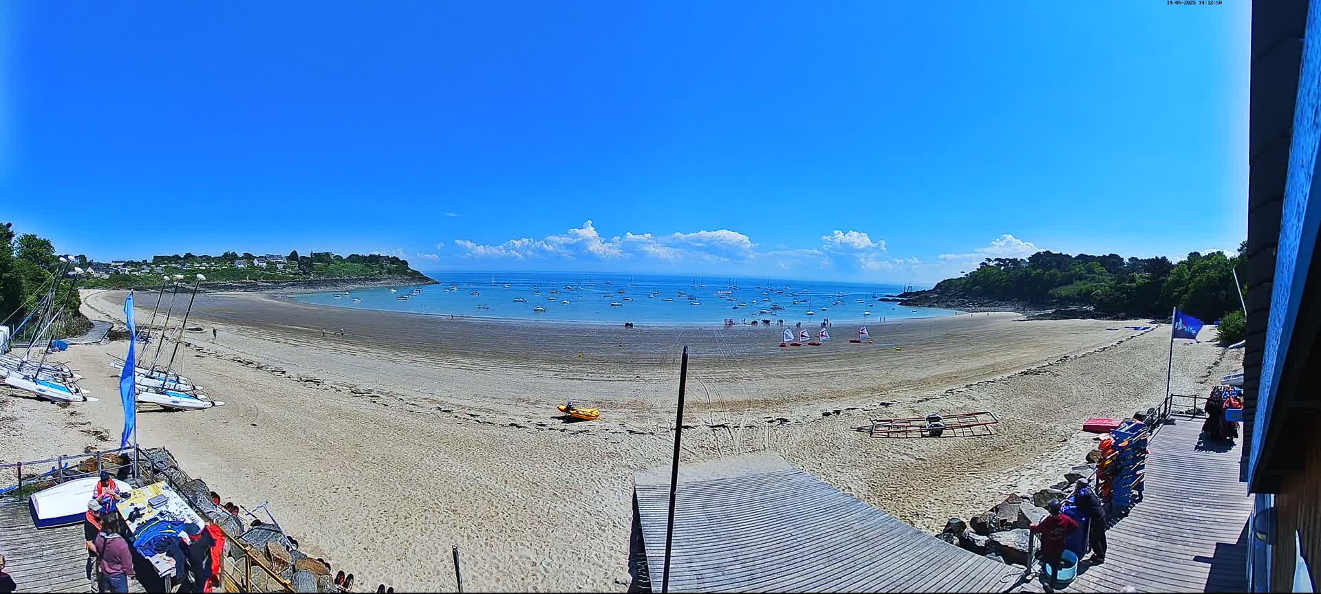 A sandy beach curves around a calm bay filled with sailboats under a mostly sunny sky with some scattered clouds.