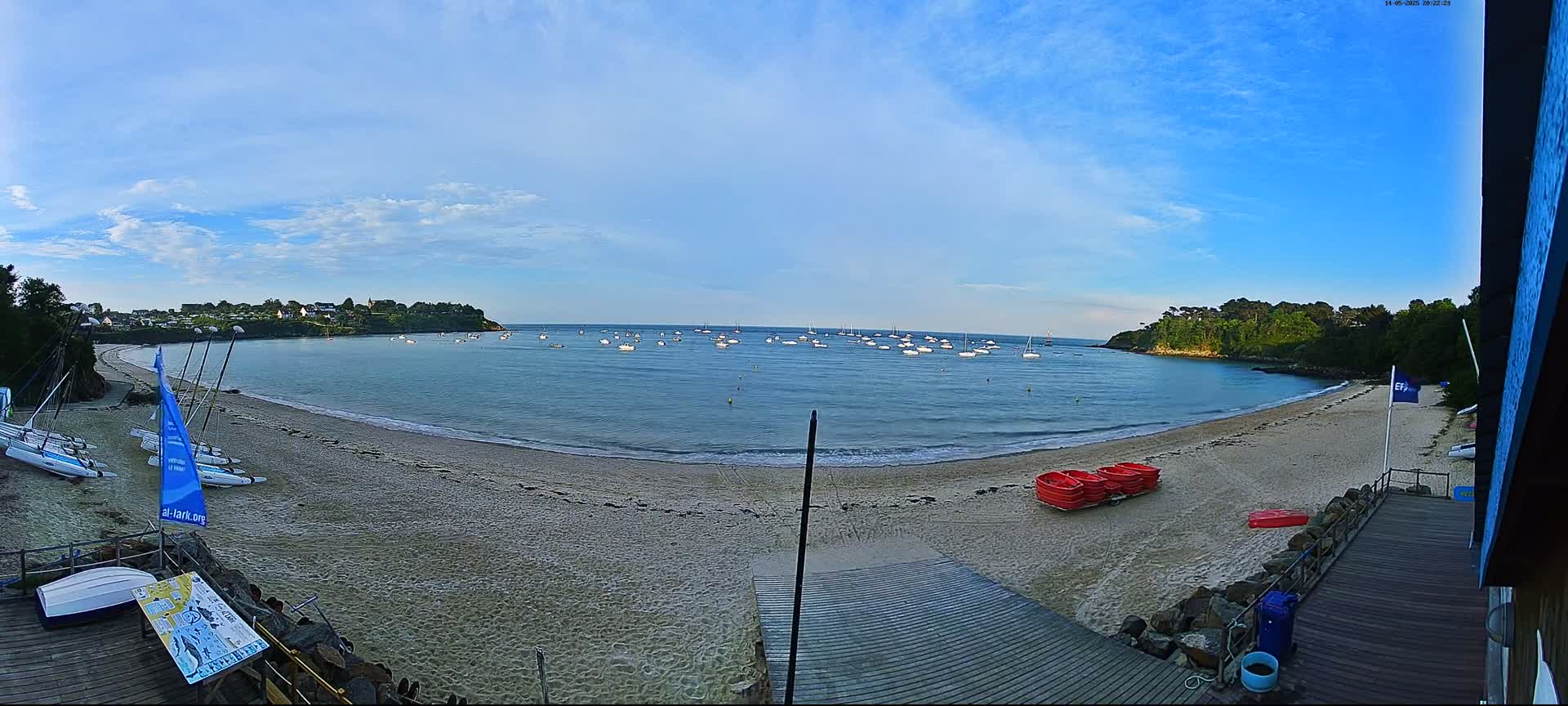 A sandy beach curves around a calm bay under a mostly clear, blue sky, with numerous small boats visible on the water and sailboats on the shore.