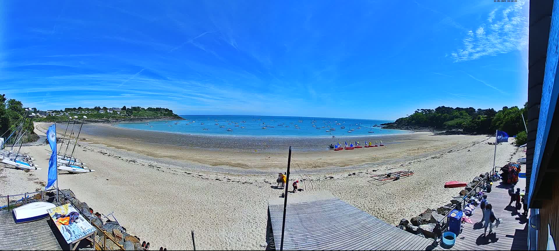 A sandy beach with sailboats in the water is viewed from a wooden boardwalk on a sunny day with a mostly clear blue sky.