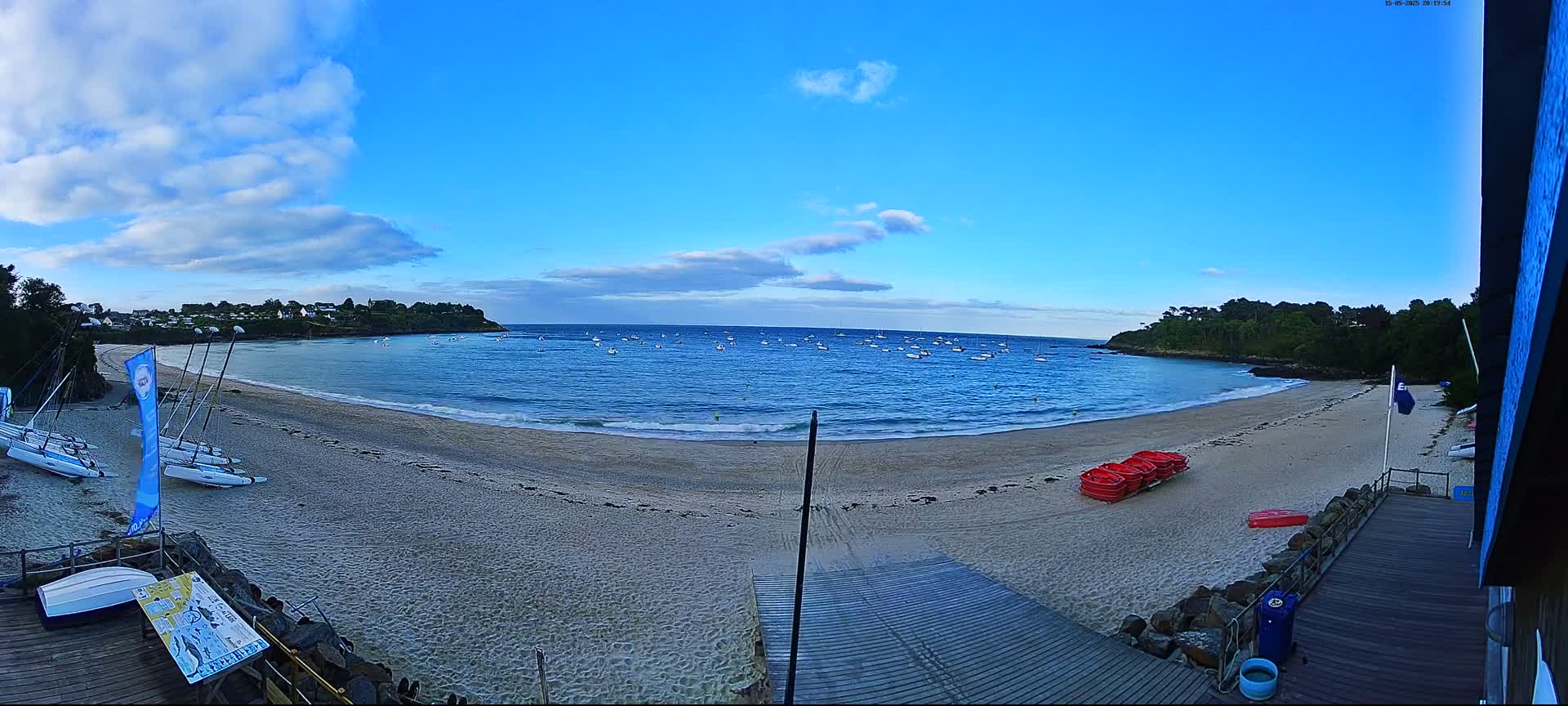 A sandy beach with calm, blue water, numerous small boats anchored offshore, and several sailboats and red rowboats on the shore under a partly cloudy sky.