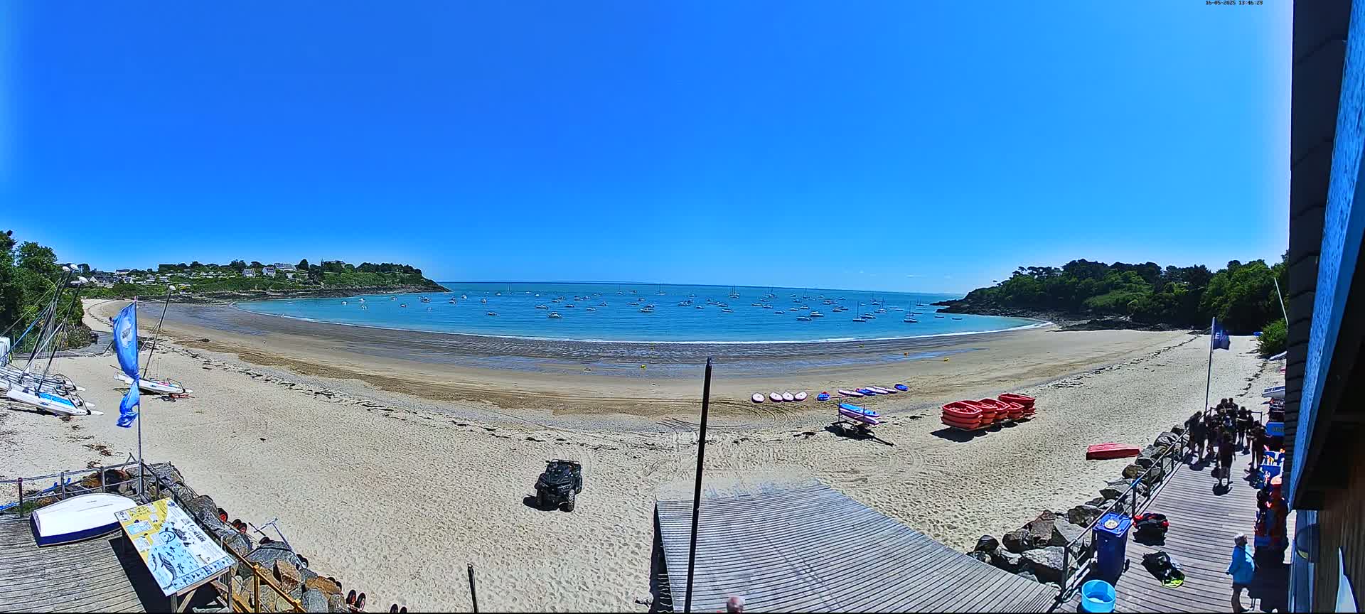 A sandy beach curves around a calm, blue bay dotted with sailboats under a clear, sunny sky.
