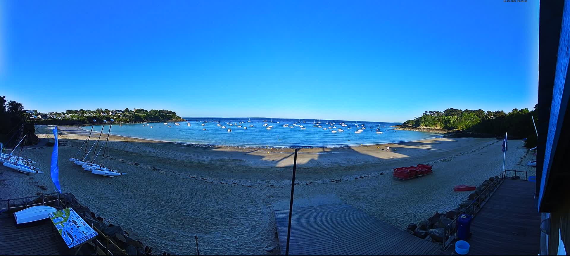 A sandy beach with several sailboats in the calm water, under a clear blue sky.