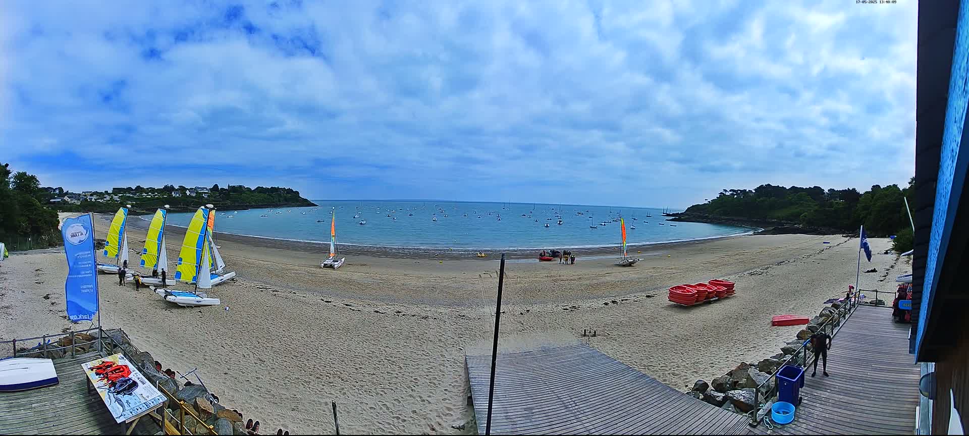 A sandy beach with several sailboats on the shore and many more in the calm bay under a partly cloudy sky.