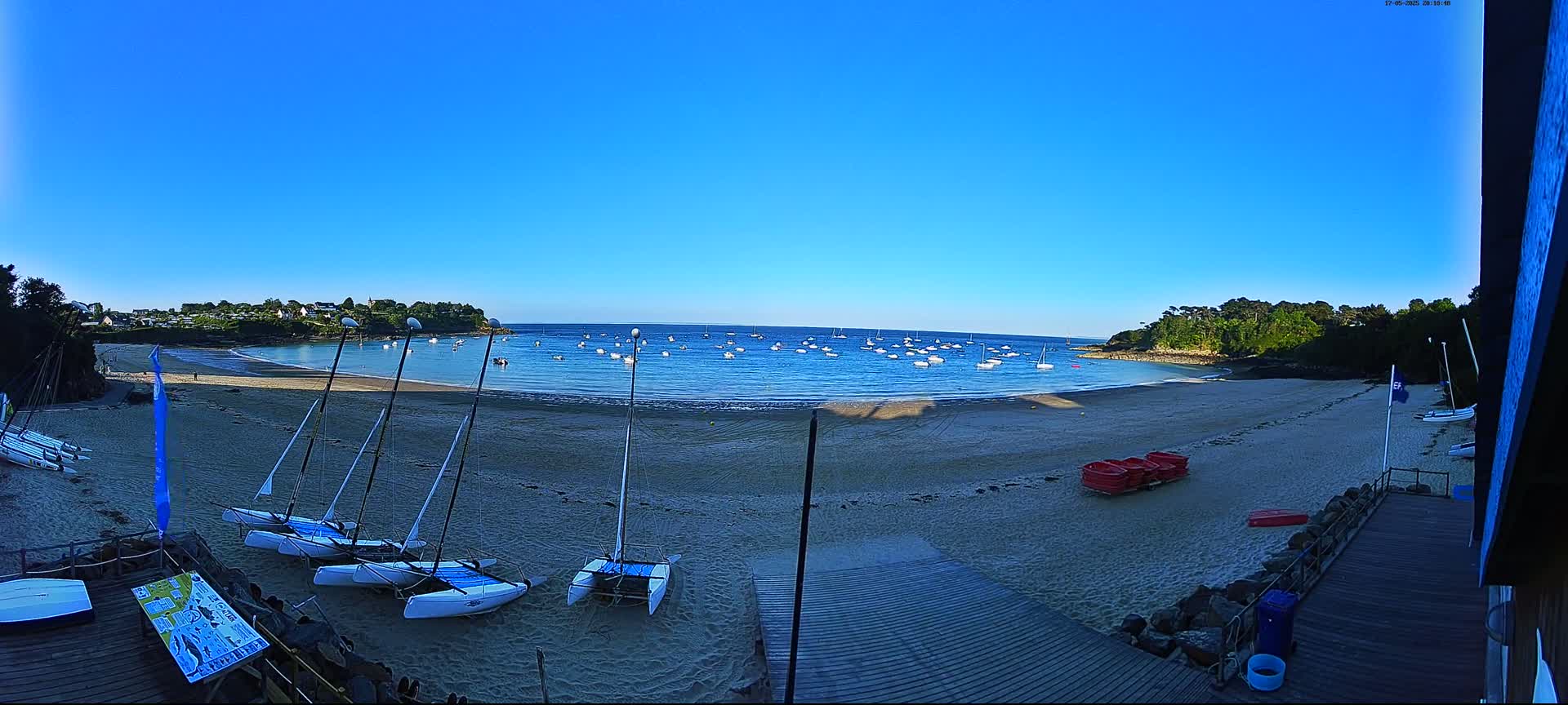 A sandy beach with several sailboats on the shore and many more in the calm blue water, under a clear blue sky.
