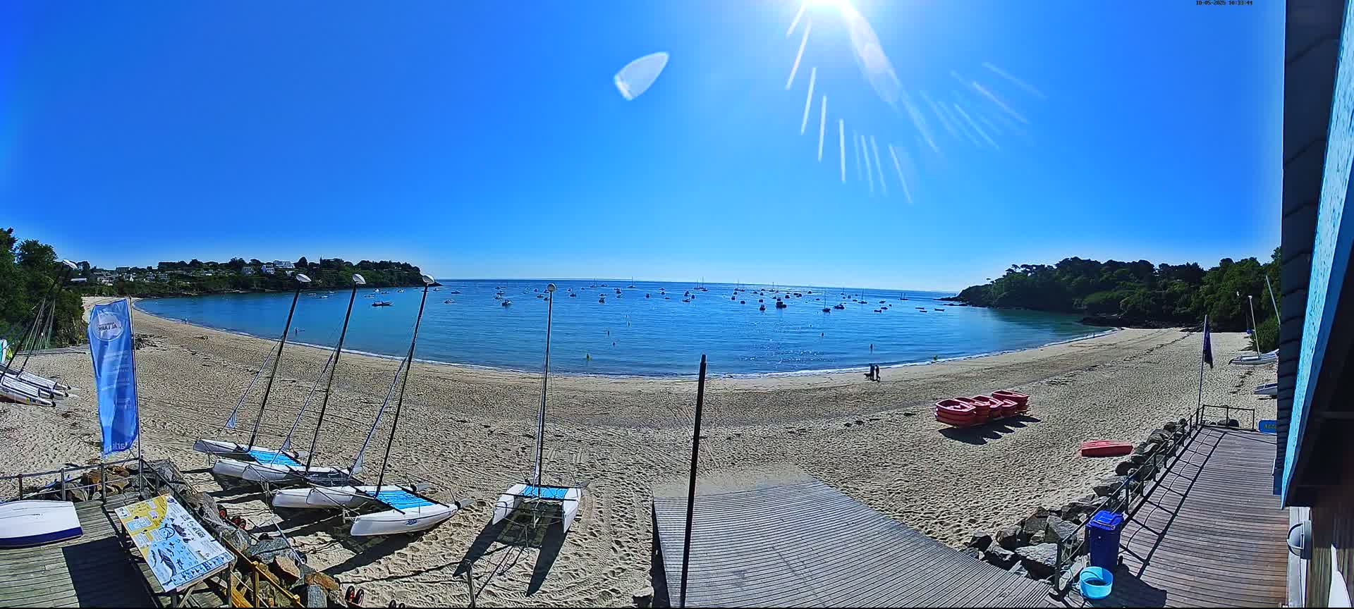 A sandy beach with several sailboats on the shore and many more in the calm, blue water under a bright, sunny sky.