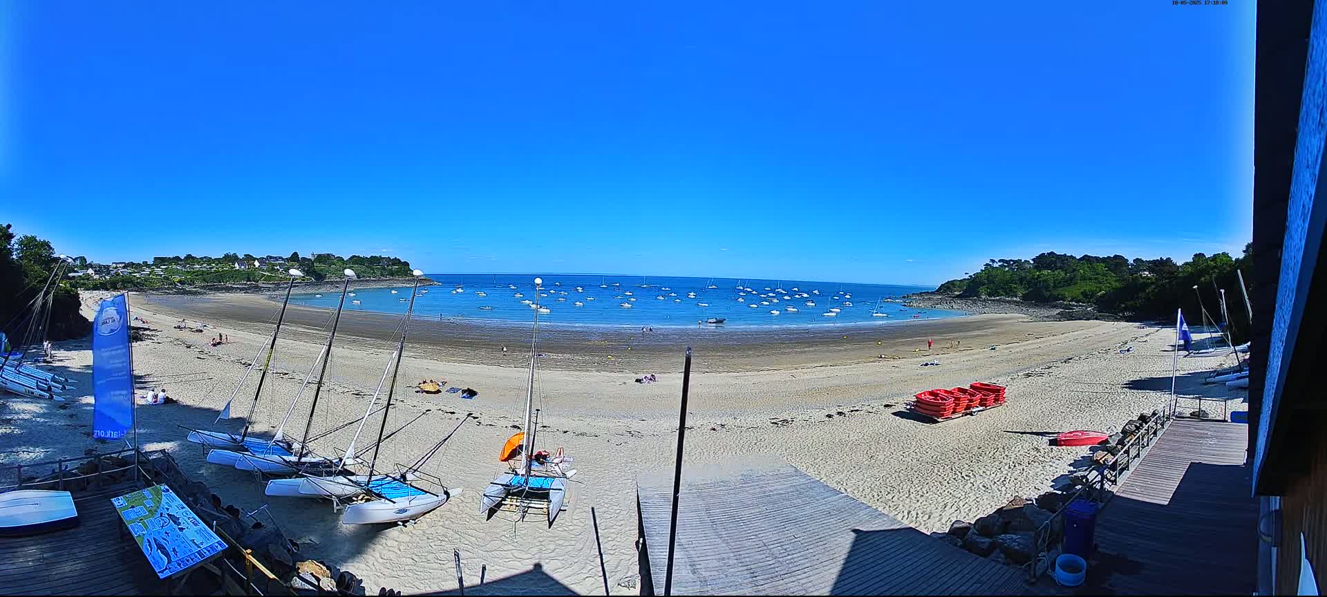 A sandy beach with numerous sailboats in the water and several catamarans on the shore under a clear blue sky.