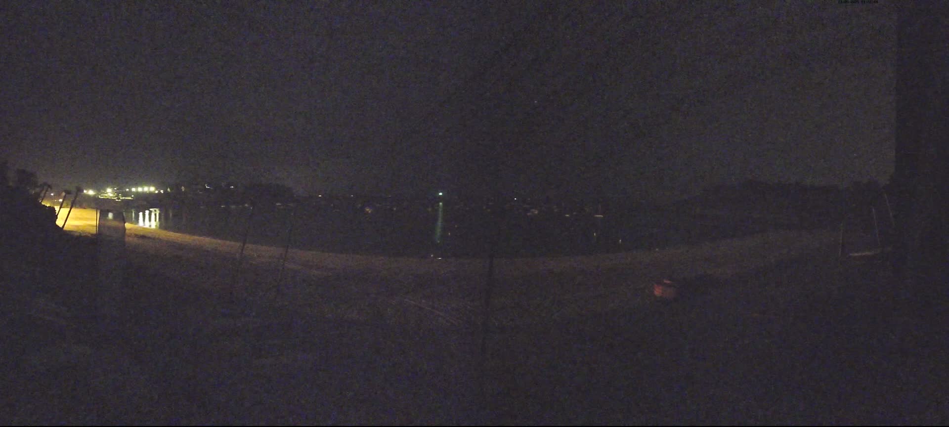 At night, dimly lit lights are visible in the distance across a dark body of water, with a dark, empty foreground.
