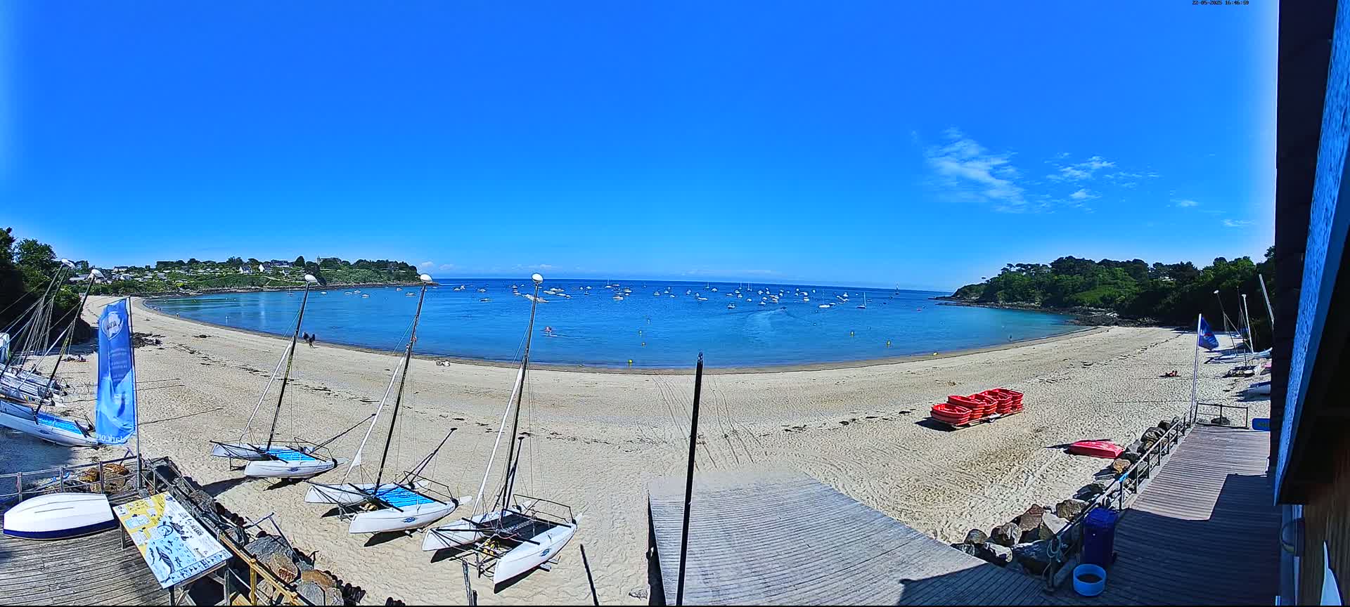 A sandy beach curves around a calm, blue bay dotted with sailboats under a clear, sunny sky.
