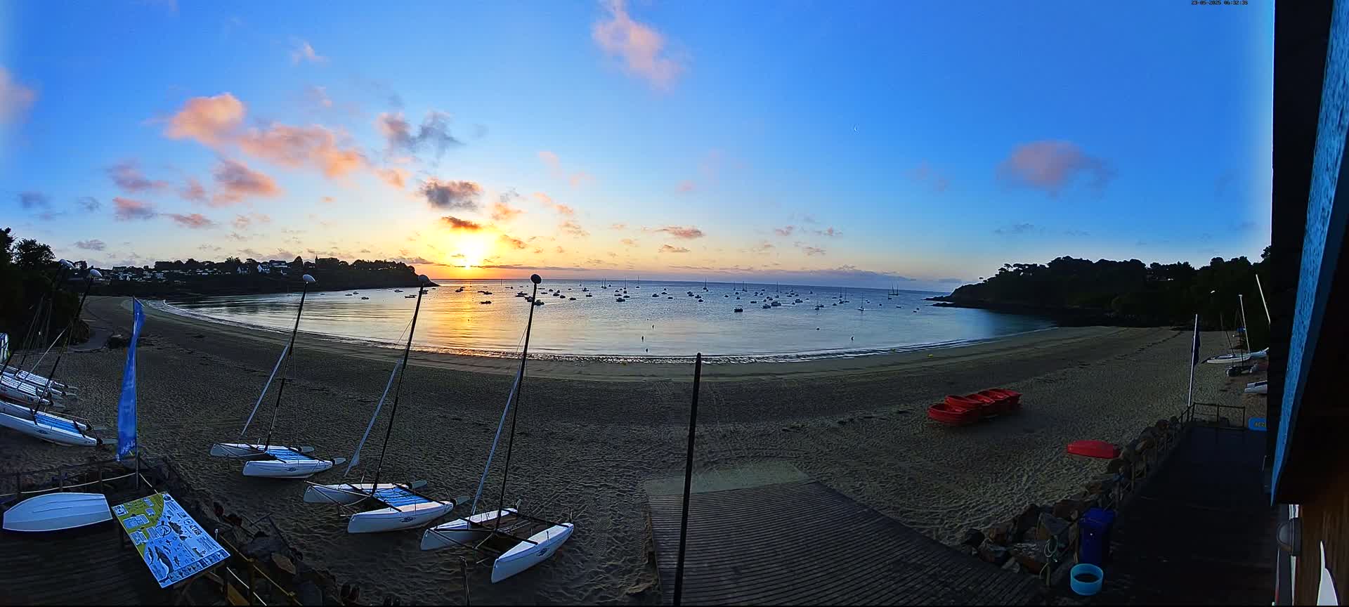 A sandy beach at sunrise, with several sailboats on the calm water and more sailboats beached on the sand.