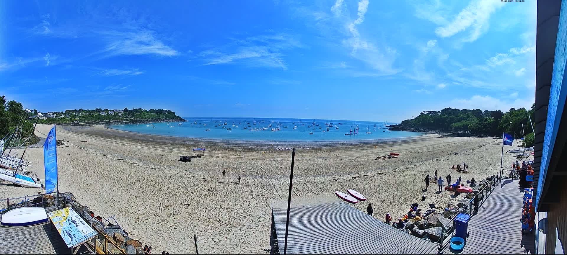 A sandy beach curves around a calm bay under a bright, sunny sky, with numerous sailboats on the water and people relaxing on the sand.