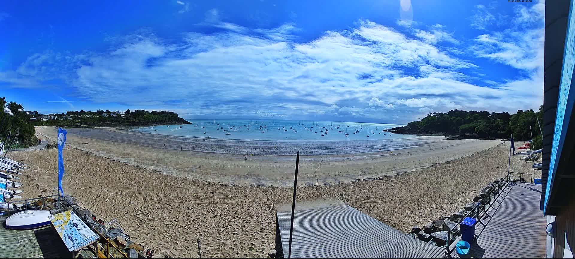 A sandy beach curves around a calm bay under a mostly sunny sky with scattered clouds, numerous small boats are visible in the water.