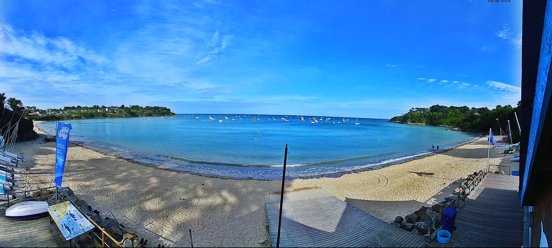 A sandy beach curves around a calm, blue bay dotted with sailboats under a bright, sunny sky.