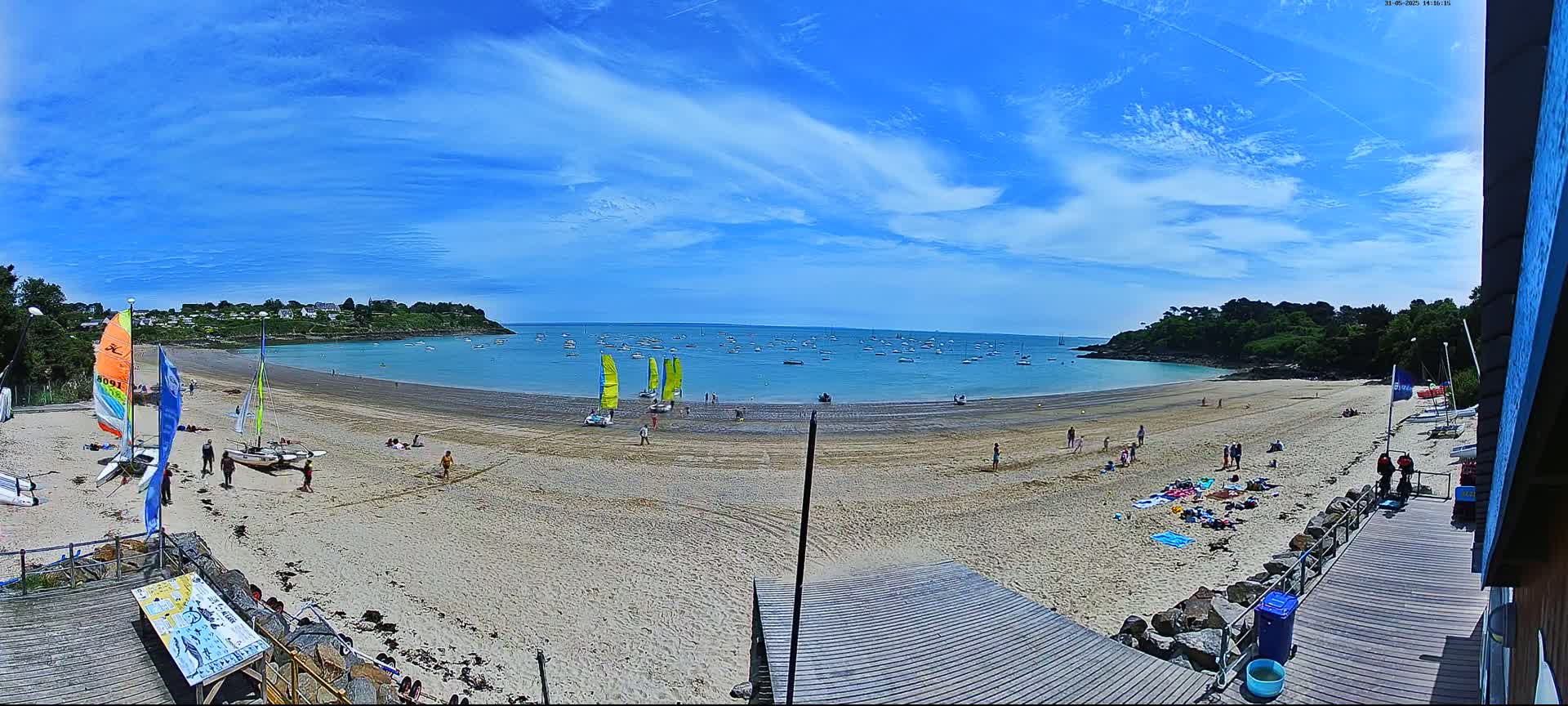 A sandy beach with people sunbathing and several sailboats in the calm water under a mostly sunny sky.