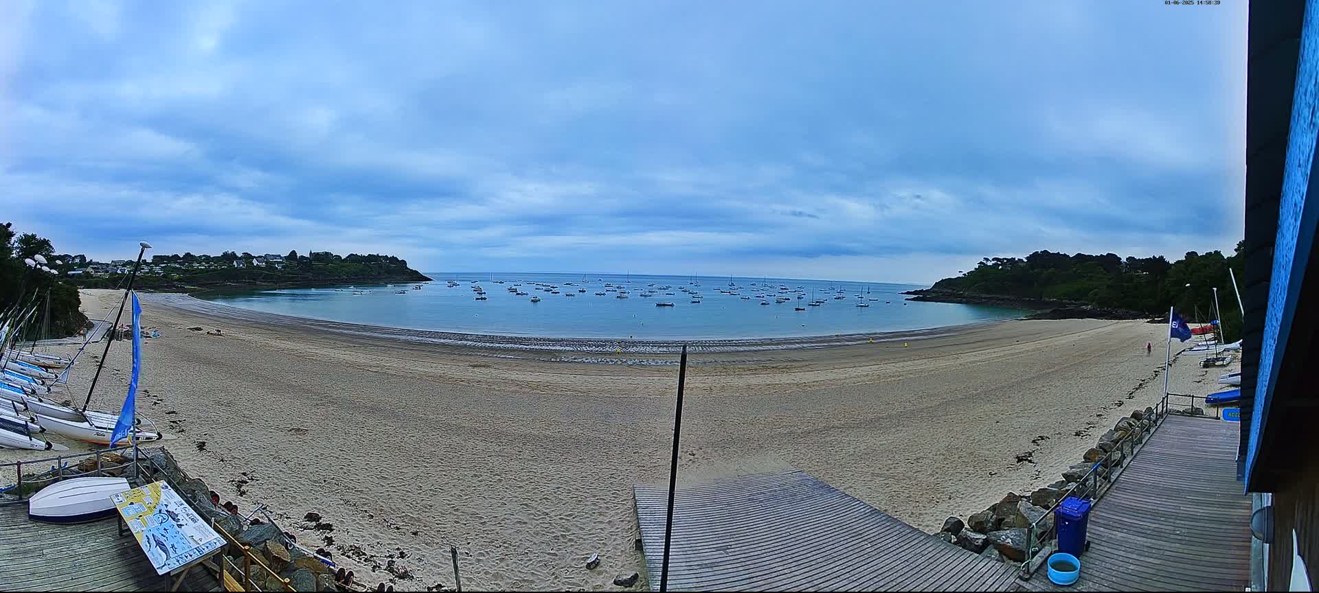 A sandy beach curves around a calm bay dotted with numerous small boats under a mostly cloudy sky.