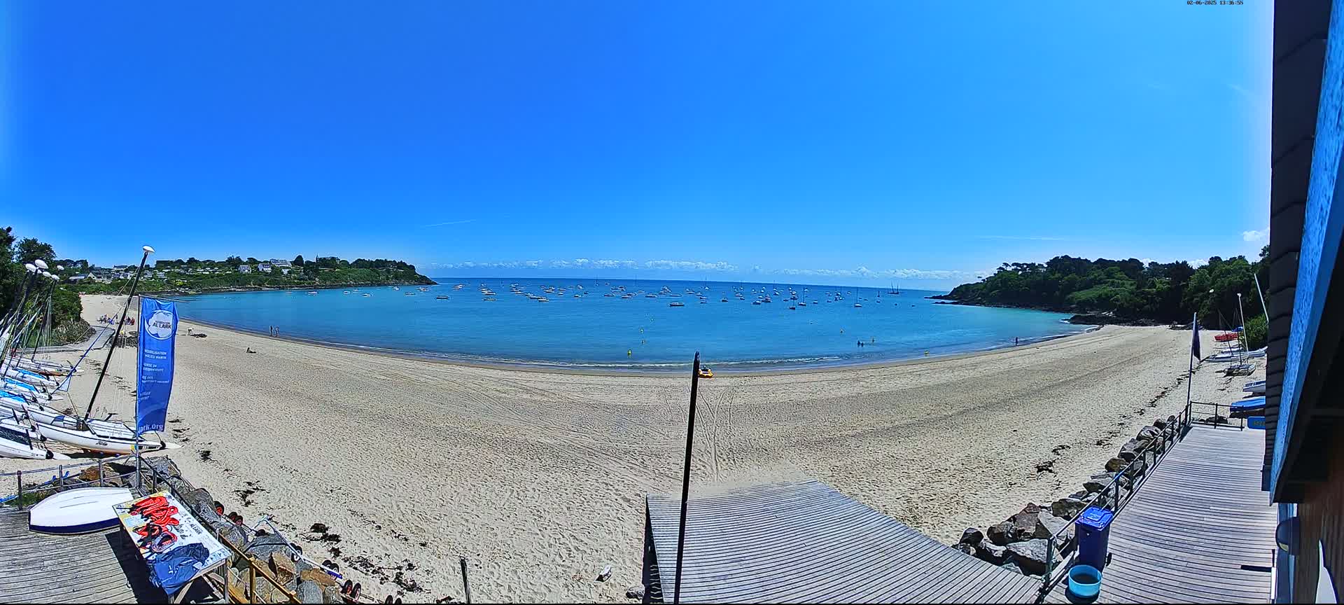 A sandy beach curves around a calm, blue bay dotted with sailboats under a clear, sunny sky.